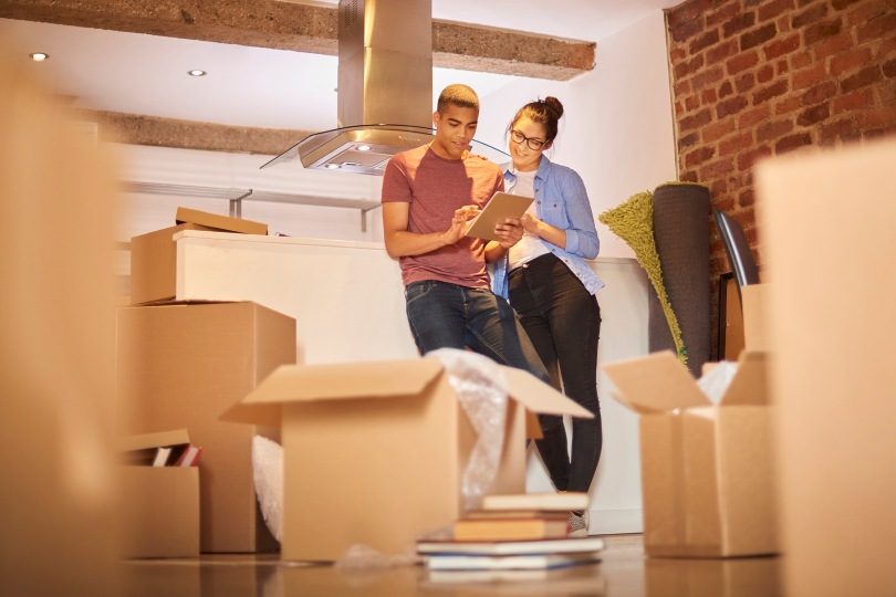 Young couple researches homes on a tablet while surrounded by moving boxes